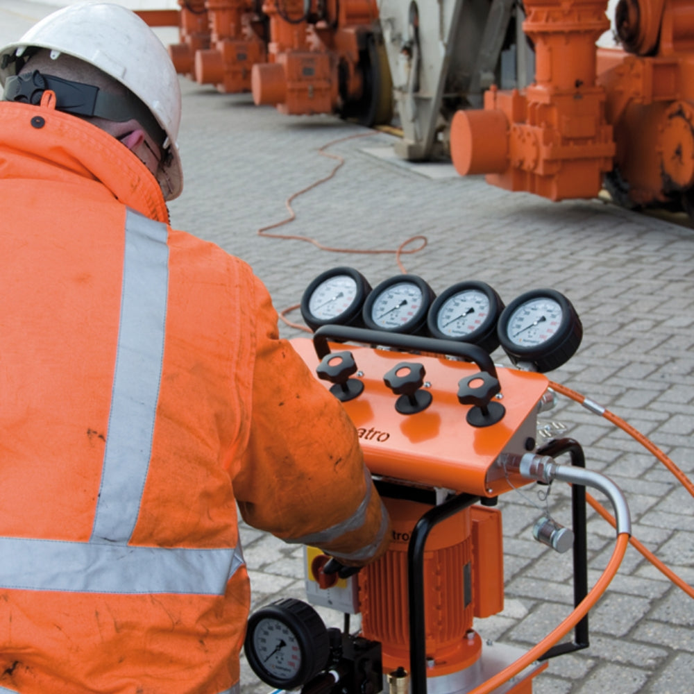 Arbejder i orange betjener Holmatro Manifold med fire manometre og ventiler på en flisebelagt overflade.