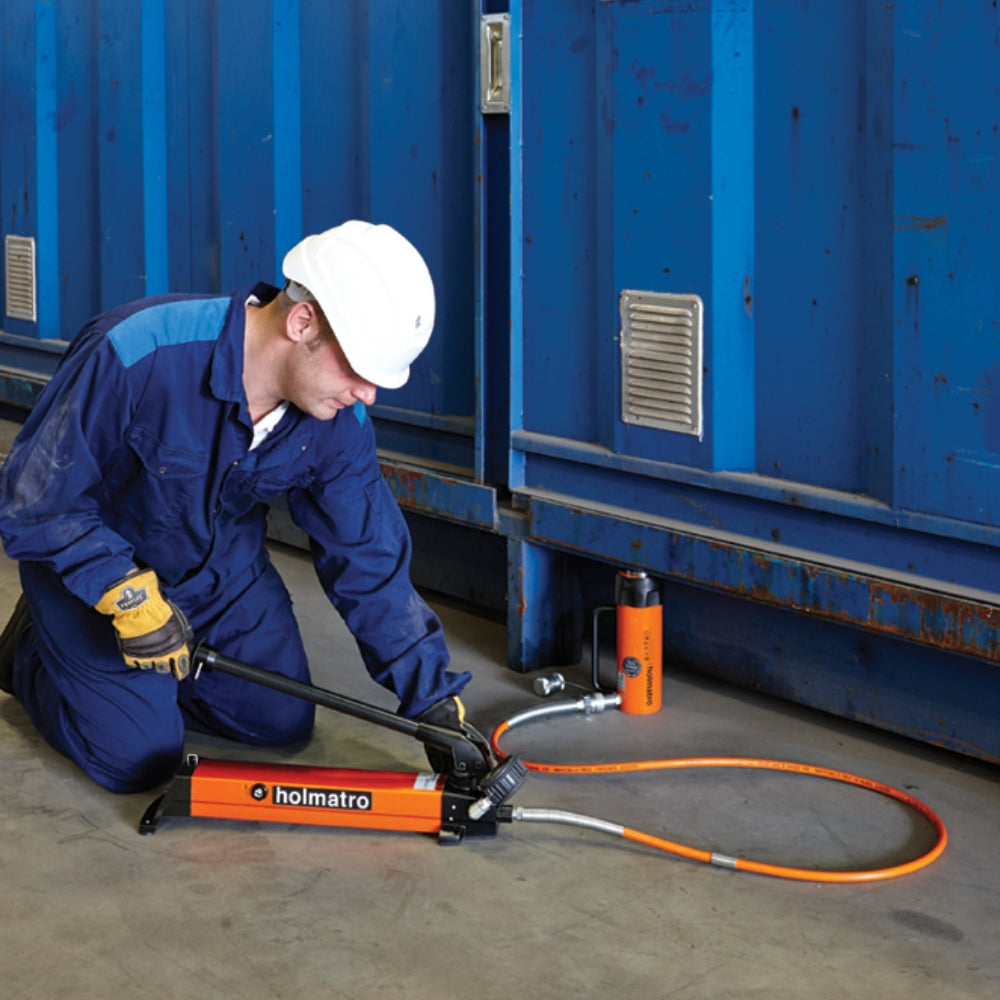 Arbejder iført hjelm og handsker bruger orange Holmatro håndpumpe foran blå container.