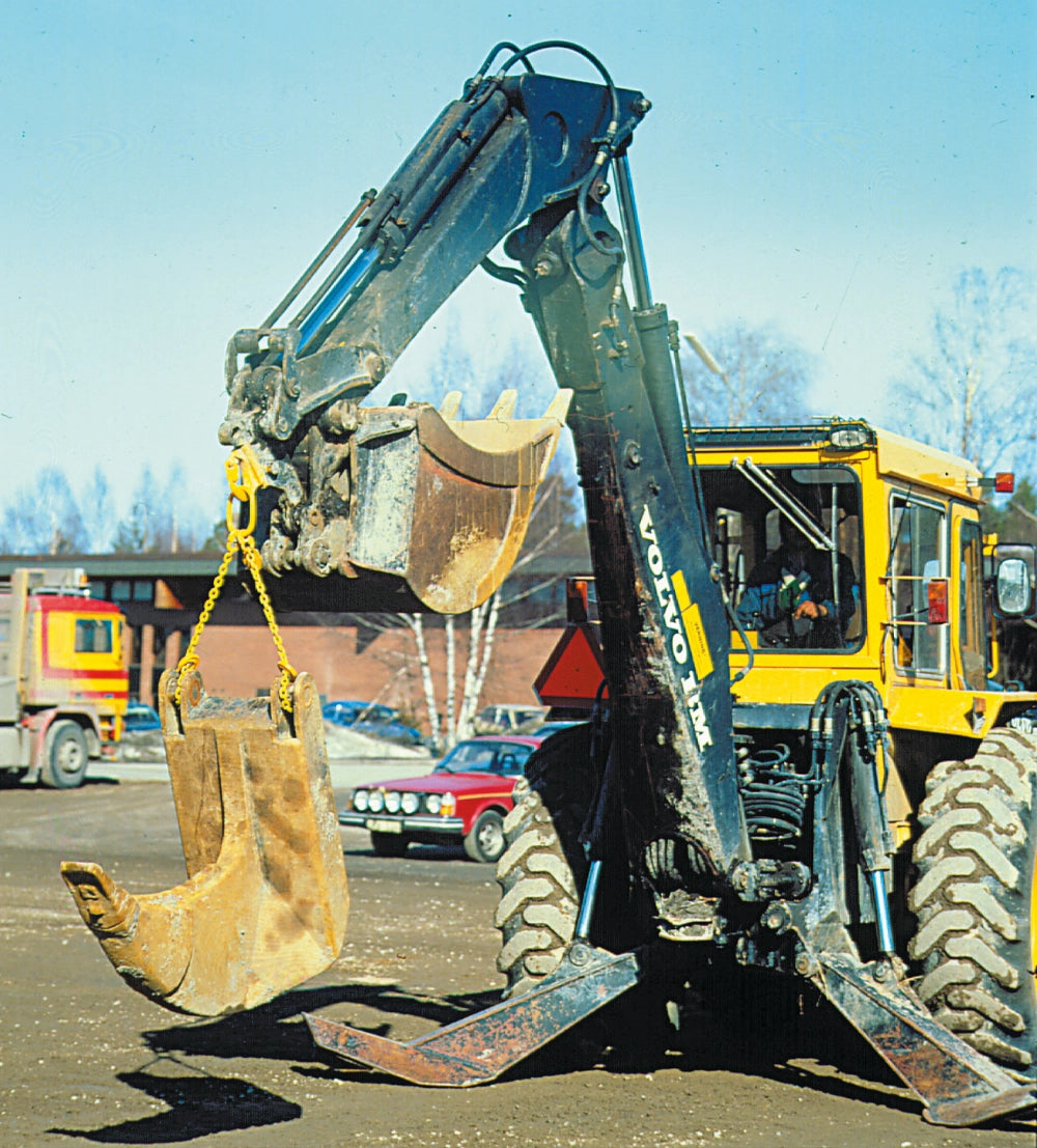 Gravemaskine løfter tungt objekt med gul Gunnebo-krog, baggrund med lastbil og biler.