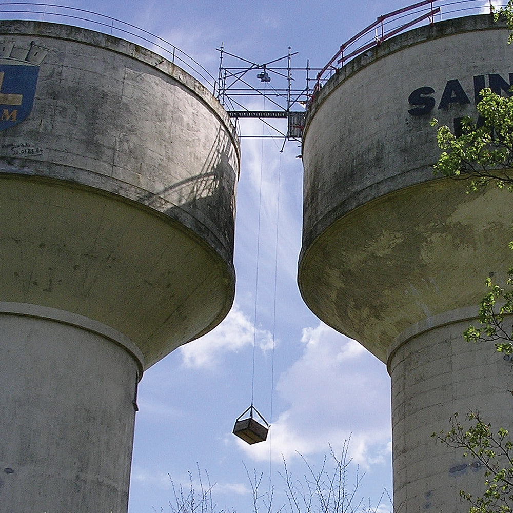 Platform hænger mellem to store betonstrukturer, understøttet af wire, med blå himmel og skyer.
