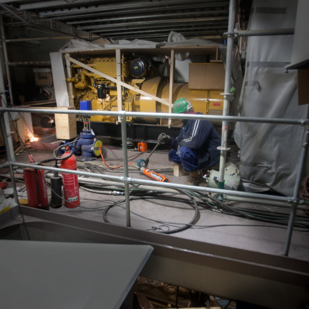 Person bruger orange Holmatro donkraft ved maskineri i industriel omgivelser med sikkerhedsudstyr.