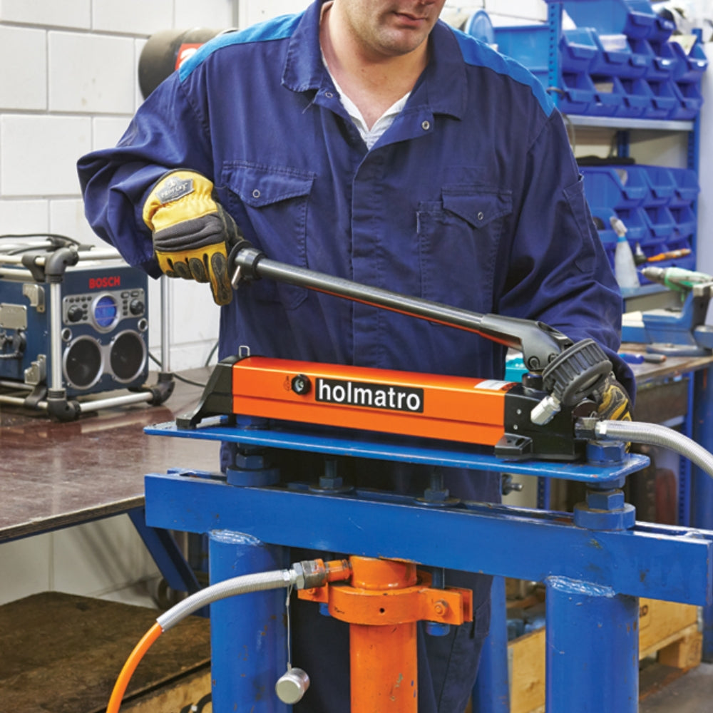 Person i arbejdstøj bruger orange Holmatro håndpumpe i værksted. Mærke tydeligt.